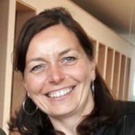 Smiling woman with dark hair and hoop earrings standing in a warmly lit indoor corridor.